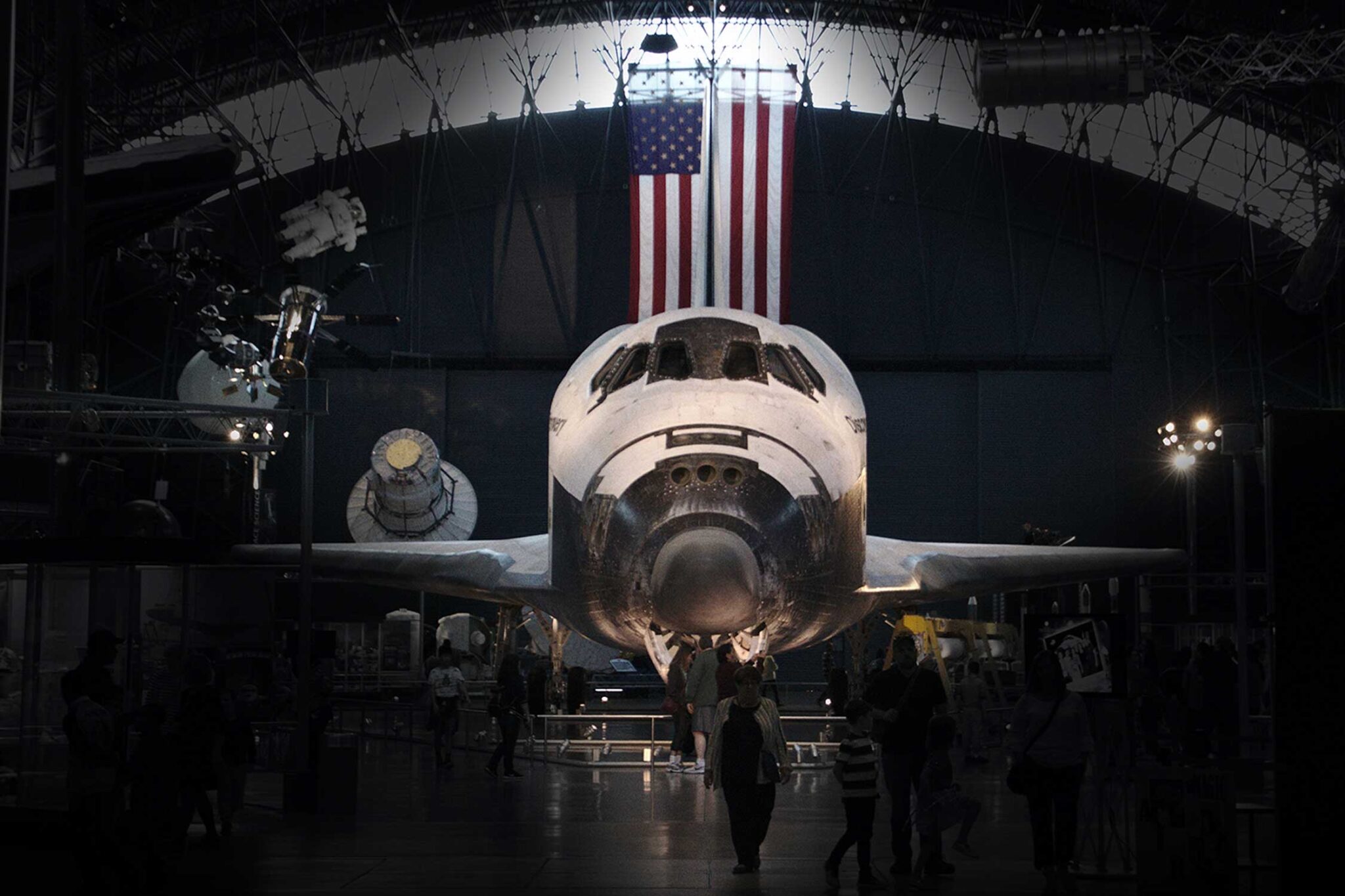 a space aircraft sits in a dark room with an American flag hanging behind it