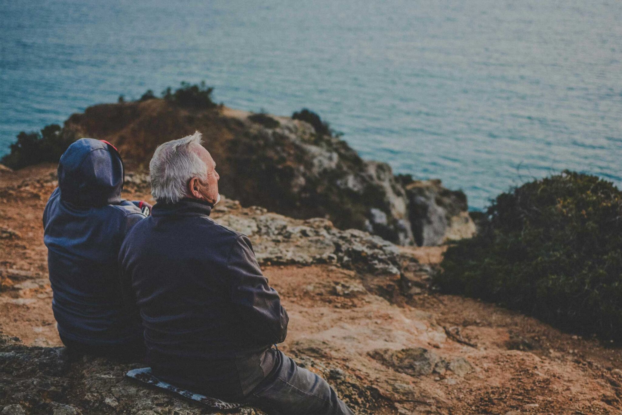 an older couple sits on a rocky hill, staring at a large body of water